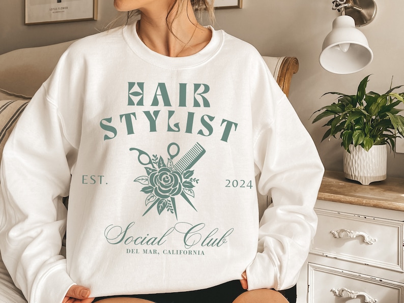 K&ouml;nnte beinhalten: Wei&szlig;er Pullover mit einem gr&uuml;nen Grafikdesign, auf dem "Hair Stylist" mit einer Rose, einer Schere und einem Kamm steht. Der Text darunter lautet "Social Club" und "Del Mar, Kalifornien".