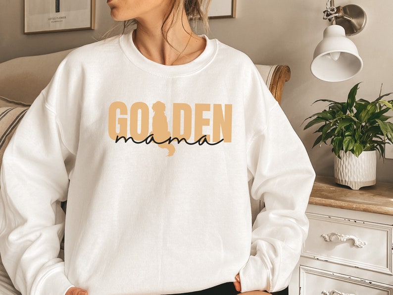 May include: White sweatshirt with the text "GOLDEN mama" in a tan color with a silhouette of a golden retriever in the "O".