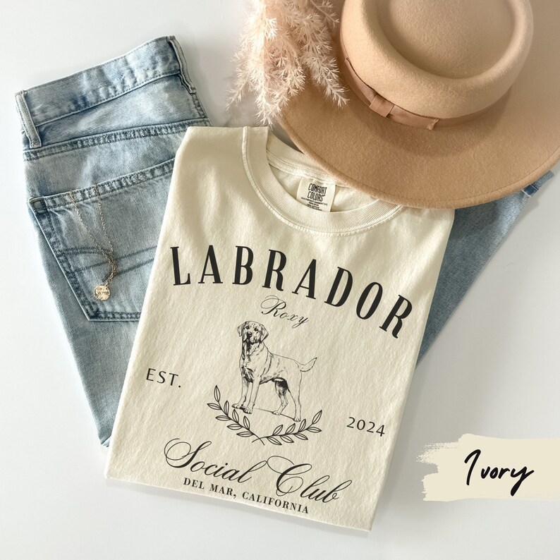 May include: Ivory t-shirt with a black graphic design featuring a Labrador Retriever, the text "Labrador Social Club", and the words "Roxy", "Est. 2024", and "Del Mar, California".
