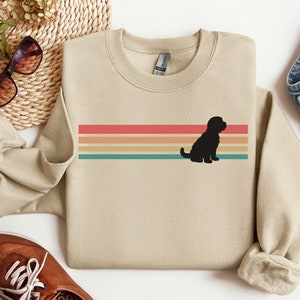 May include: A beige sweatshirt with a retro rainbow stripe design and a black silhouette of a dog sitting in front of the stripes.