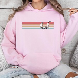 May include: A light pink hoodie featuring a retro design with a horizontal striped background in red, orange, teal, and tan. The design includes two Siamese cats. The hoodie has a front pocket and a drawstring hood.