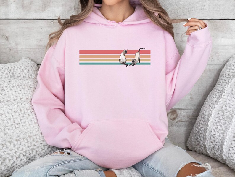 May include: A light pink hoodie featuring a retro design with a horizontal striped background in red, orange, teal, and tan. The design includes two Siamese cats. The hoodie has a front pocket and a drawstring hood.