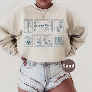 May include: A sand-colored sweatshirt with a blue stamp design featuring medical-themed illustrations and the text "X-ray Tech." The design includes images of a skeleton, hand, and other medical symbols. The sweatshirt is paired with denim shorts.