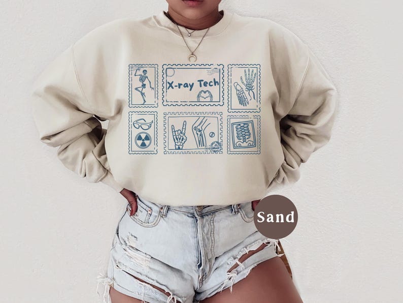 May include: A sand-colored sweatshirt with a blue stamp design featuring medical-themed illustrations and the text "X-ray Tech." The design includes images of a skeleton, hand, and other medical symbols. The sweatshirt is paired with denim shorts.