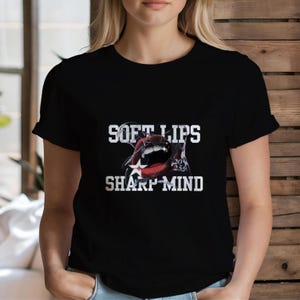以下が含まれることがあります： 白い歯と星がついた赤い開いた口のグラフィックがプリントされた黒いTシャツ。Tシャツには「Soft Lips Sharp Mind」というテキストが白でプリントされています。