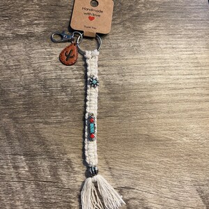 May include: White macrame keychain with a turquoise and red beaded charm, a silver star charm, and a brown leather cactus charm. The keychain has a silver ring and a silver lobster clasp. The tag attached to the keychain reads "Handmade with love. Thank you."