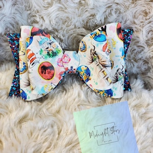 May include: A large, colorful bow with a white background and a variety of images, including a hat, a globe, and a building. The bow is decorated with blue and black glitter. The bow is on a white fluffy surface. The text "Midnight Stars by Les" is on a blue and green gradient background.