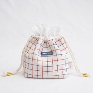 Puede incluir: Una bolsa de cordón blanco con un patrón a cuadros rojo y azul y un volante blanco en la parte superior. La bolsa tiene una etiqueta azul marino que dice "Sewing Stroll".