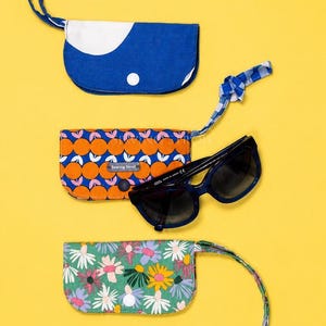 May include: Three fabric pouches with wrist straps and a pair of dark blue sunglasses. One pouch is solid blue with a white accent, one features an orange citrus pattern with a Sewing Stroll label, and one has a colorful floral print on green.