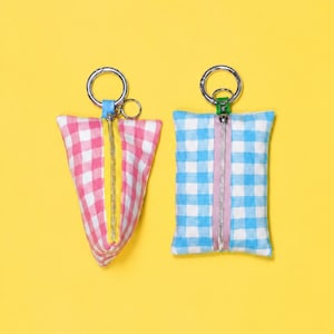 Peut inclure: Deux petites pochettes en tissu avec des anneaux porte-clés argentés. Une pochette est triangulaire avec un motif vichy rose et blanc et une fermeture éclair jaune. L'autre est rectangulaire avec un motif vichy bleu et blanc et une fermeture éclair lavande.