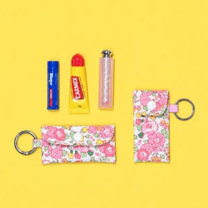 May include: Two rectangular fabric pouches with a pink and green floral pattern, each featuring a metal keyring and a white snap closure. Beside them are a blue Blistex tube, a yellow Carmex tube, and a clear, cylindrical cosmetic container.