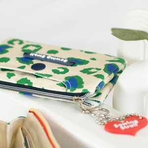 May include: A beige zippered pouch with a green and blue leopard print design. The pouch has a black button and a zipper pull with the text "Home Buddies". A red heart-shaped keychain with the text "Smiling Buddies" is attached to the zipper pull.