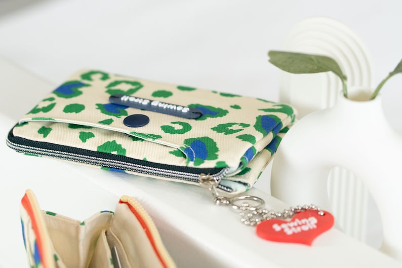 May include: A beige zippered pouch with a green and blue leopard print design. The pouch has a black button and a zipper pull with the text "Home Buddies". A red heart-shaped keychain with the text "Smiling Buddies" is attached to the zipper pull.