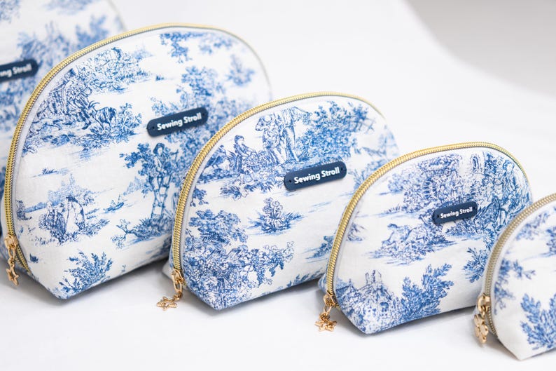 May include: A set of white and blue toiletry bags with a landscape design. The bags have gold zippers and a small blue tag that reads "Sewing Stroll". The bags are in various sizes.