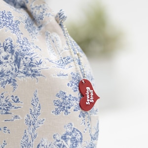 May include: A white fabric bag with a blue toile pattern and a red heart-shaped tag that says "Sewing Stroll".