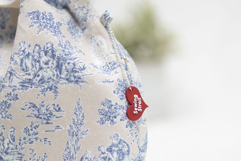 May include: A white fabric bag with a blue toile pattern and a red heart-shaped tag that says "Sewing Stroll".