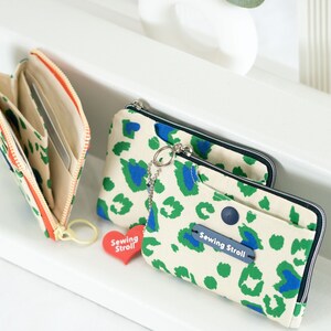 May include: Two zippered wallets with a leopard print design in shades of green, blue, and white. The wallets have a zipper closure and a small charm with the text "Sewing Stroll" on a red heart.
