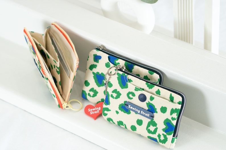 May include: Two zippered wallets with a leopard print design in shades of green, blue, and white. The wallets have a zipper closure and a small charm with the text "Sewing Stroll" on a red heart.