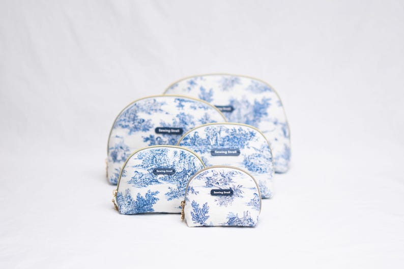 May include: A set of five cosmetic bags in varying sizes, featuring a blue and white toile pattern. Each bag has a gold zipper and a small rectangular label that reads "Sewing Street". The bags are arranged on a white background.