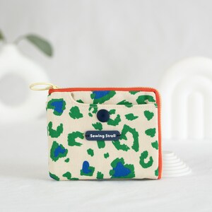 May include: A small, cream-colored zippered pouch with a green and blue leopard print design. The pouch has a red zipper and a blue button closure. The text "Sewing Stroll!" is printed on a blue label.