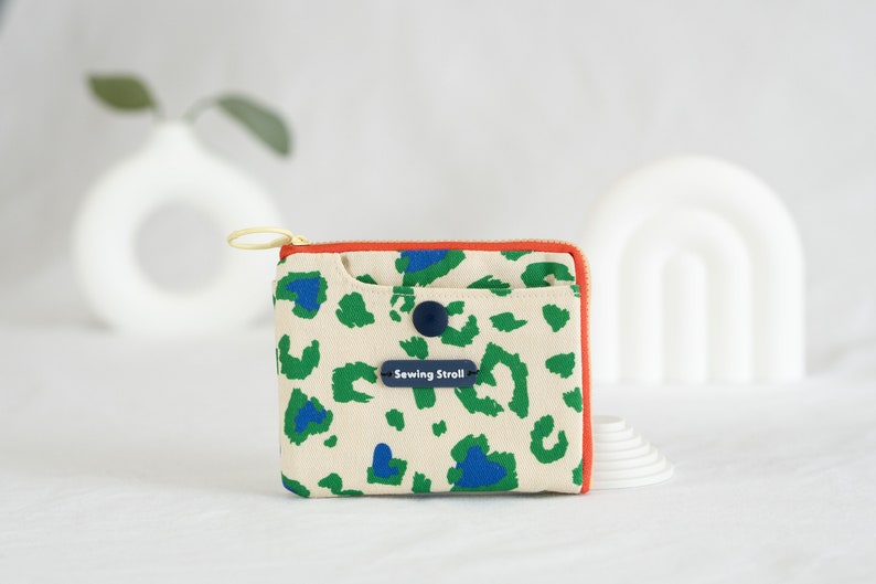 May include: A small, cream-colored zippered pouch with a green and blue leopard print design. The pouch has a red zipper and a blue button closure. The text "Sewing Stroll!" is printed on a blue label.