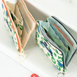 May include: Two open zippered wallets with a leopard print design. One wallet is beige with orange zipper and the other is beige with green and blue accents. Both wallets have a key ring attached.