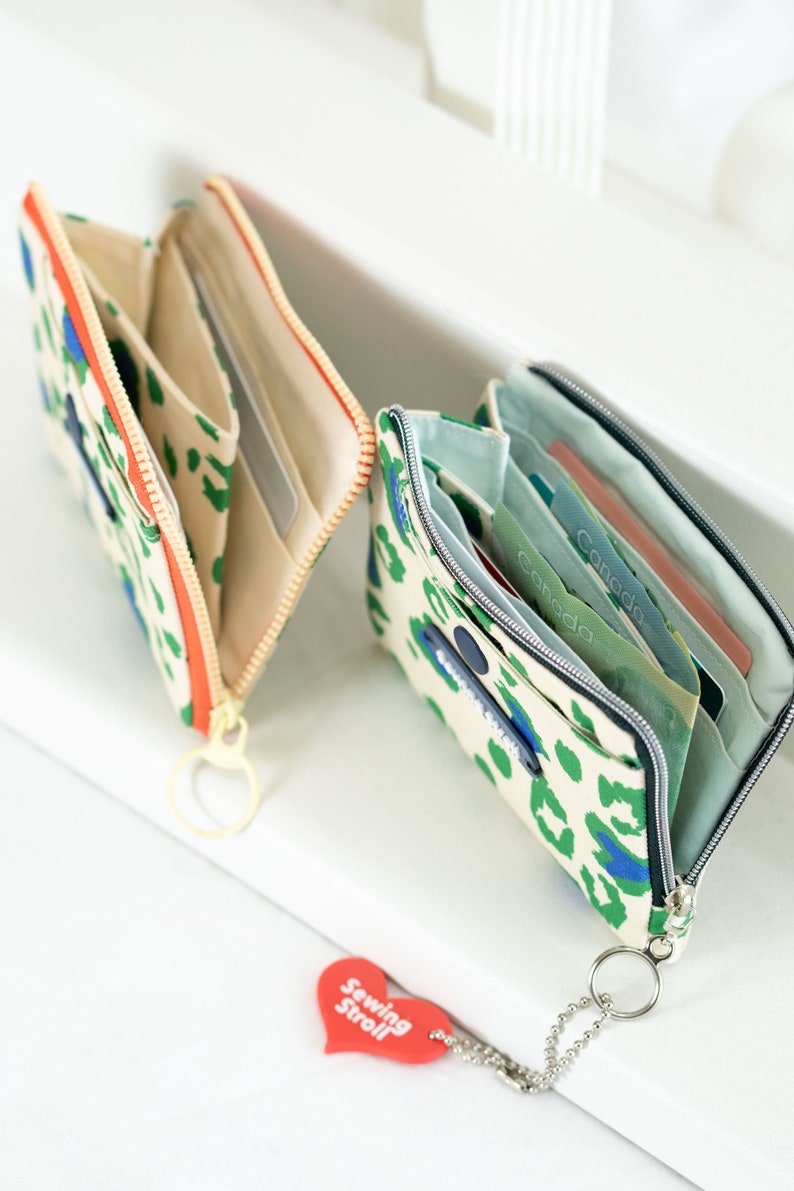 May include: Two open zippered wallets with a leopard print design. One wallet is beige with orange zipper and the other is beige with green and blue accents. Both wallets have a key ring attached.
