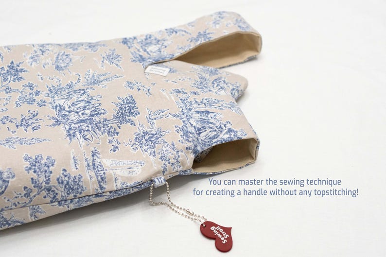 May include: A beige fabric tote bag with a blue and white floral pattern. The bag has a handle that is sewn into the fabric without any topstitching. A small red heart-shaped tag with the text "Just Sew" is attached to the bag.
