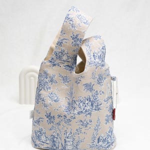 May include: A beige tote bag with a blue and white toile pattern. The bag has a single handle and a small red tag attached to it.