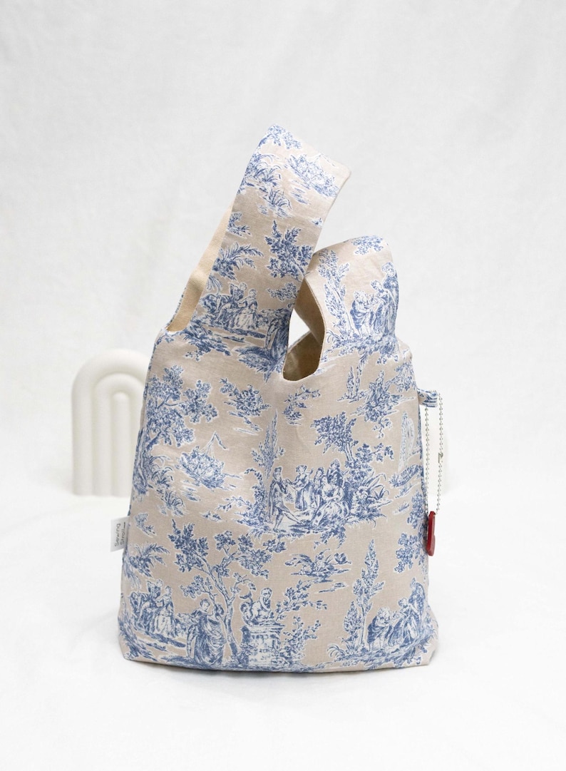 May include: A beige tote bag with a blue and white toile pattern. The bag has a single handle and a small red tag attached to it.