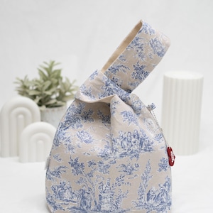 May include: A beige fabric tote bag with a blue and white toile pattern. The bag has a knotted handle and a small red tag with the word "Studio" on it.