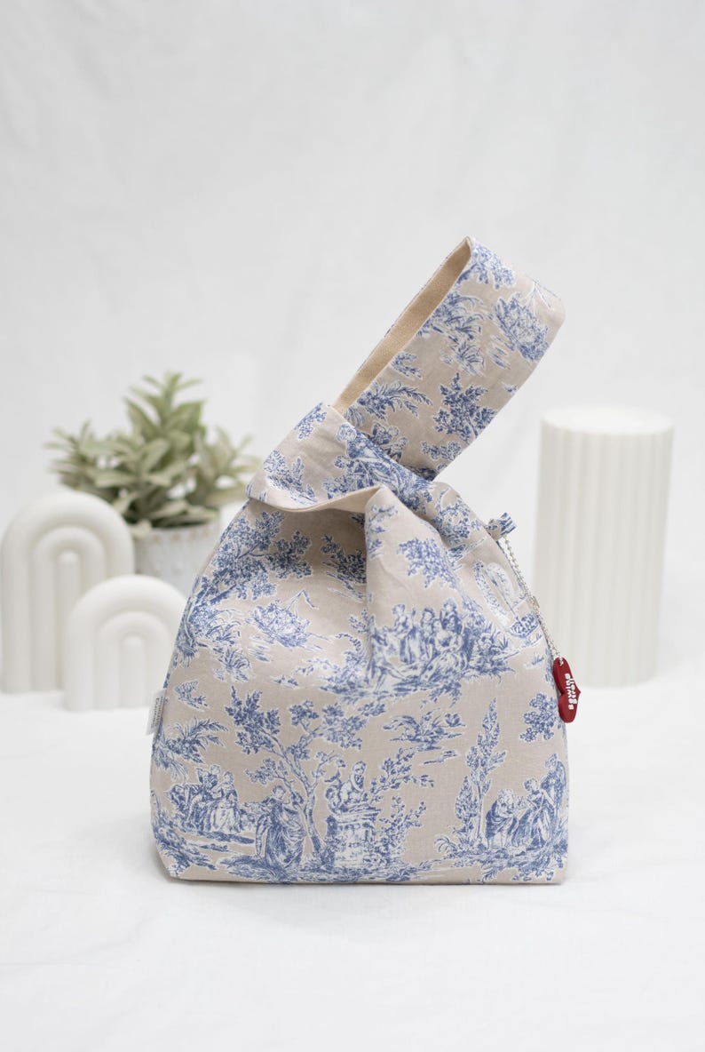 May include: A beige fabric tote bag with a blue and white toile pattern. The bag has a knotted handle and a small red tag with the word "Studio" on it.