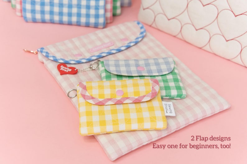 Puede incluir: Conjunto de bolsos de tela de cuadros vichy en amarillo, verde y azul, con cierres de solapa, dispuestos sobre un fondo de cuadros vichy rosa. Un charm rojo en forma de coraz&oacute;n cuelga de uno de los bolsos.