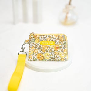 Puede incluir: Una peque&ntilde;a bolsa de cremallera floral amarilla con una correa amarilla y un cierre plateado. La bolsa tiene una etiqueta amarilla que dice "Sewing Stroll".