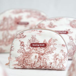 May include: A set of three white cosmetic bags with a red toile pattern featuring a landscape design. Each bag has a half-moon shape with a zipper closure and a small rectangular tag that reads "Sewing Stroll".