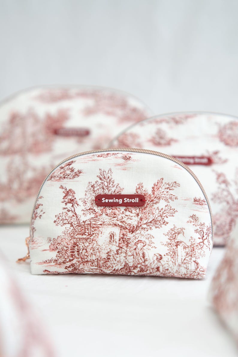 May include: A set of three white cosmetic bags with a red toile pattern featuring a landscape design. Each bag has a half-moon shape with a zipper closure and a small rectangular tag that reads "Sewing Stroll".