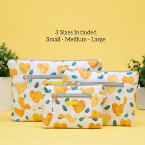 May include: Set of four zippered pouches in various sizes, featuring a white background with a repeating pattern of yellow hearts and teal leaf shapes. The pouches have silver zippers and a yellow label.