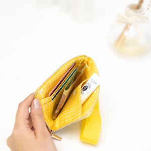 Puede incluir: Una bolsa amarilla con cremallera, con un patr&oacute;n de lunares blancos y una correa amarilla. La bolsa est&aacute; abierta y contiene varias tarjetas y un peque&ntilde;o tubo de crema de manos.