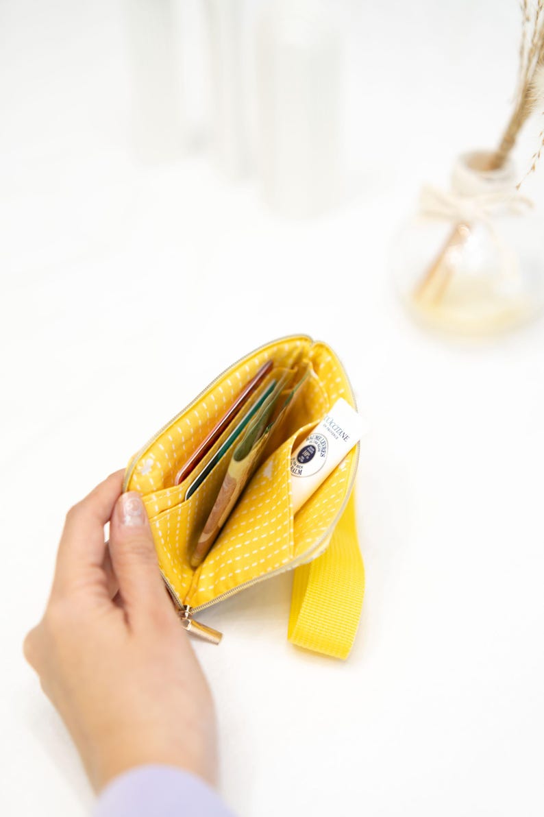 Puede incluir: Una bolsa amarilla con cremallera, con un patr&oacute;n de lunares blancos y una correa amarilla. La bolsa est&aacute; abierta y contiene varias tarjetas y un peque&ntilde;o tubo de crema de manos.