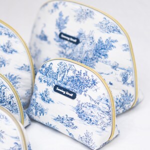 May include: Several white and blue toiletry bags with gold zippers are arranged on a white surface. The bags feature a blue and white pastoral scene print. Each bag has a small blue tag that reads "Sewing Stroll".