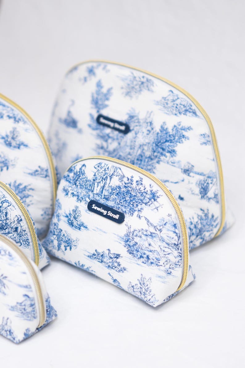 May include: Several white and blue toiletry bags with gold zippers are arranged on a white surface. The bags feature a blue and white pastoral scene print. Each bag has a small blue tag that reads "Sewing Stroll".
