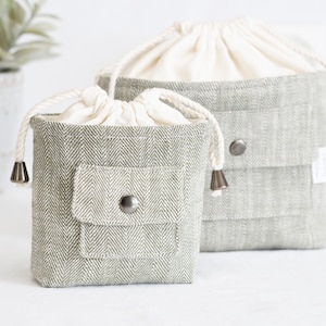 May include: Two grey linen drawstring bags with white cotton lining. The smaller bag has a front pocket with a button closure. Both bags have a white drawstring.