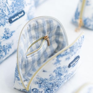 May include: Open cosmetic bag with a blue and white toile pattern, featuring a gingham interior and a gold zipper. The bag has a small pocket inside. The text "Sewing Seed" is on a small blue tag.