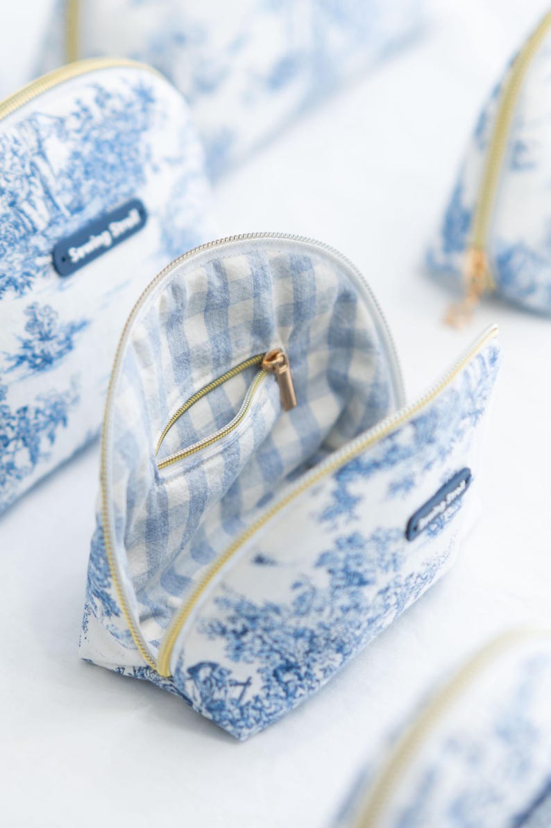 May include: Open cosmetic bag with a blue and white toile pattern, featuring a gingham interior and a gold zipper. The bag has a small pocket inside. The text "Sewing Seed" is on a small blue tag.