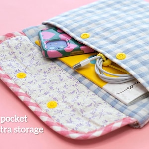 May include: Open, quilted fabric case with a blue and white gingham pattern. The case has a front pocket with a floral print and yellow snap closures. Inside, a phone with a pink and green case, a yellow notebook, and a charging cable are visible.