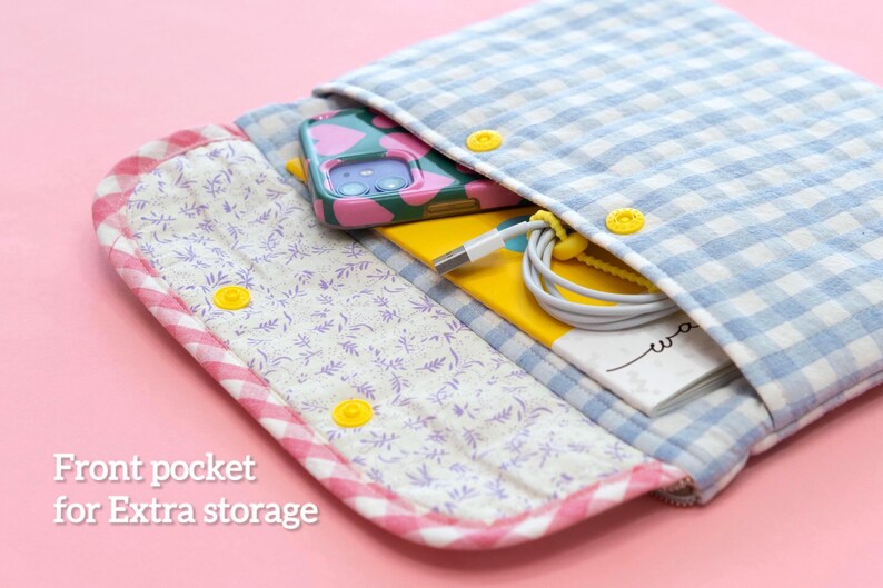 May include: Open, quilted fabric case with a blue and white gingham pattern. The case has a front pocket with a floral print and yellow snap closures. Inside, a phone with a pink and green case, a yellow notebook, and a charging cable are visible.