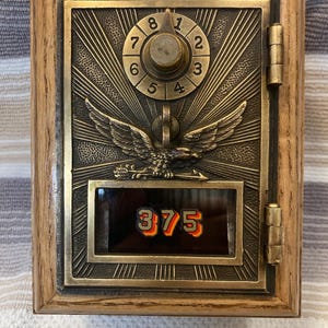 May include: A vintage-style brass post office box door mounted in a wooden frame. The door features a central eagle emblem, a circular combination dial with numbers one through eight, and a glass window displaying the black and orange numbers 375.