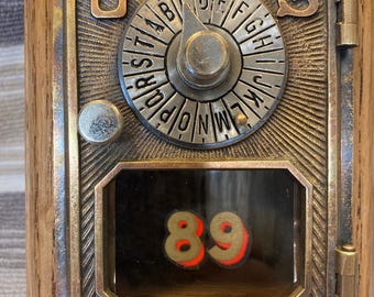 May include: A vintage-style mailbox with a wooden frame and a metal front. The metal front features a combination dial with letters and a small window displaying the number 89 in red and gold. The mailbox has the letters "U" and "S" on the top.