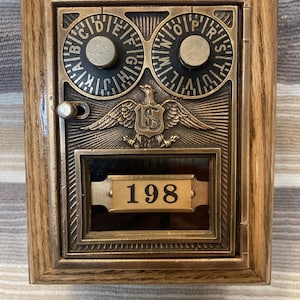May include: A vintage brass post office box door set in a wooden frame. The door features two circular combination dials with letters, a central eagle emblem with the letters US, and a rectangular glass window displaying the number 198.