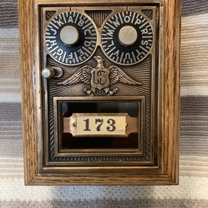 May include: A vintage-style brass post office box door mounted in a wooden frame. The door features two circular combination dials with letters, a central eagle emblem with the letters US, and a glass window displaying the number 173 on a plate.